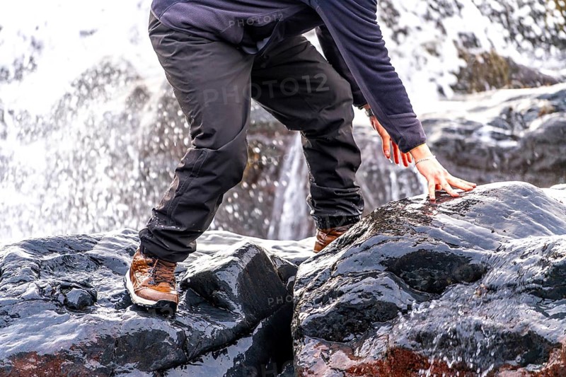 A man is standing on a rock with his hand on a wet rock. The man is wearing a black jacket and black pants. Concept of adventure and exploration