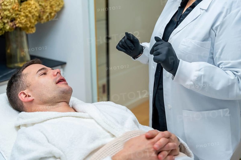 Man reclining in a robe on a treatment bed as a doctor in a lab coat and black gloves readies a cosmetic skin procedure in a clean, modern aesthetic clinic setting