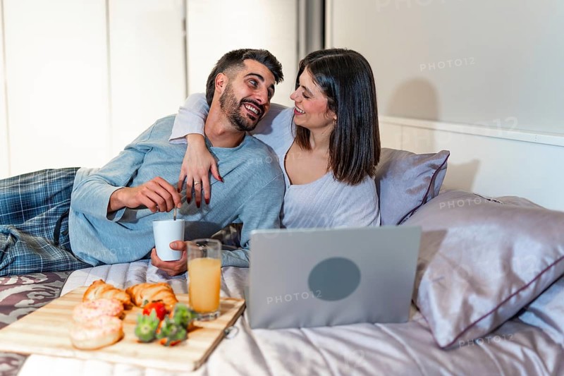 Young couple relaxing in bed, having breakfast and watching a movie on their laptop, enjoying a cozy morning together