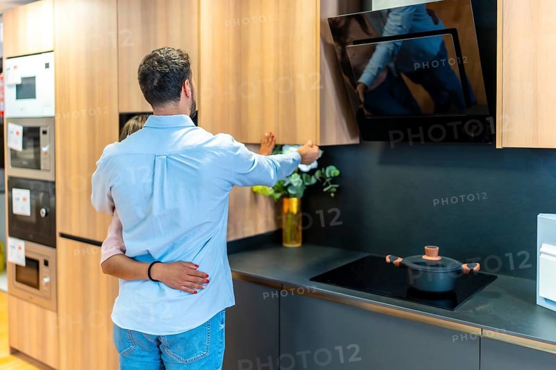 Young couple exploring kitchen design options and appliances, looking at wooden cabinets and modern cooking hoods in a showroom environment during their remodeling journey