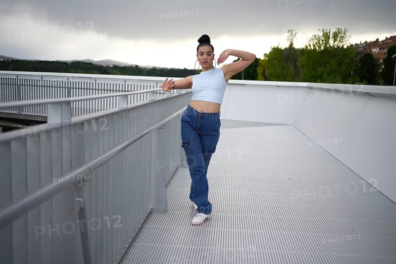A woman in blue jeans and a blue tank top is posing on a railing. The image has a moody, somewhat melancholic feel to it, as the woman is alone and isolated