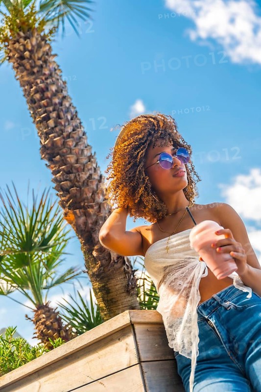 Vertical low angle view photo of a sensual latin woman with curly hair drinking milkshake standing outdoors next to palm tree in summer