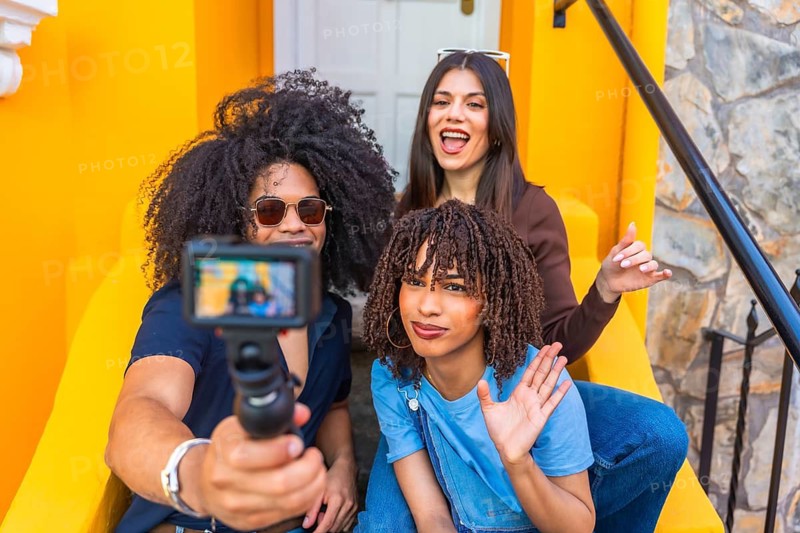 Three influencers waving and smiling at the camera while filming a vibrant video blog outdoors in an urban setting with colorful backgrounds