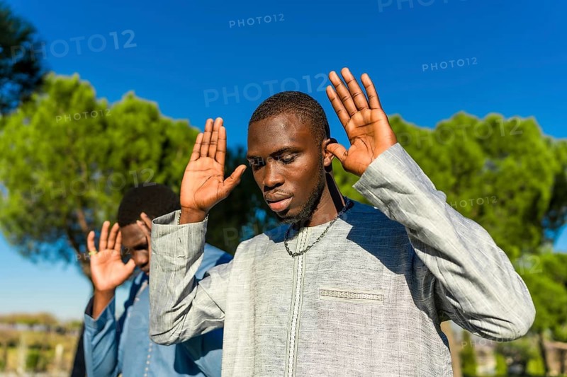 Two young senegalese men wearing traditional clothing are praying outdoors with their hands raised, possibly observing ramadan