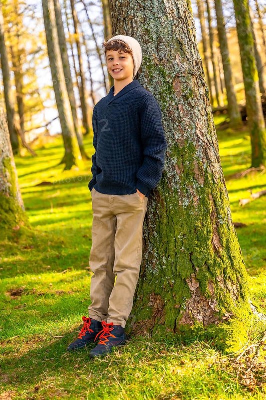 Vertical full length portrait of a caucasian happy boy with casual clothes leaning relaxed on tree trunk enjoying nature