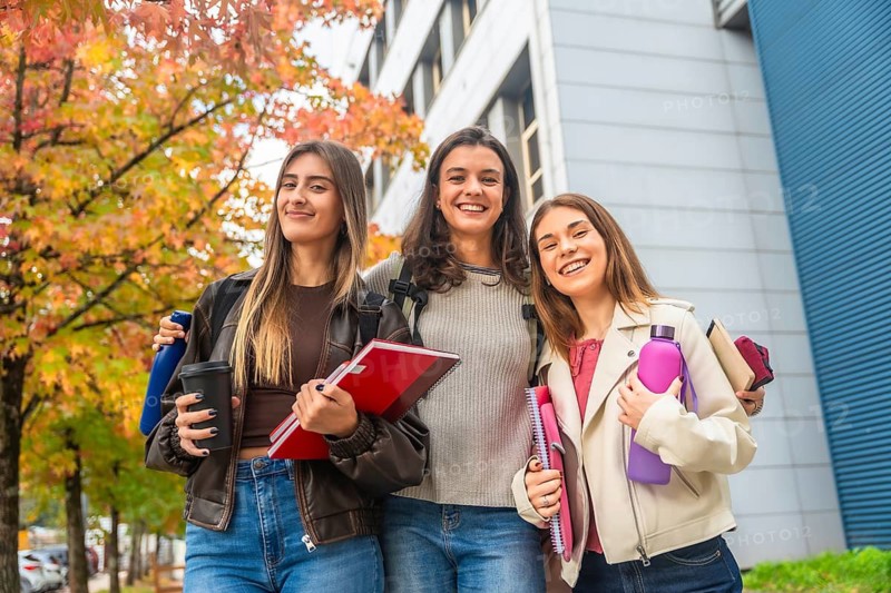 Three young women smiling and carrying notebooks, bottles, and a coffee cup, standing on a college campus with autumn trees and a modern building reflecting their joy and a back to school atmosphere