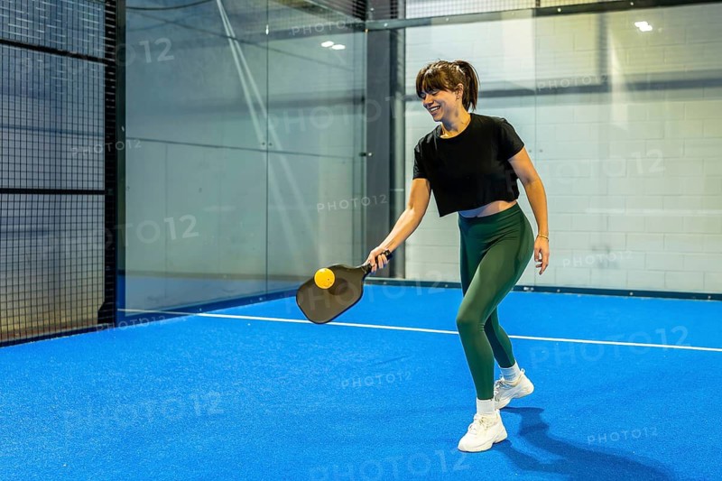 Smiling woman playing pickleball, hitting a yellow ball with a black paddle on a blue indoor court, focusing on fitness, active lifestyle, and enjoying a competitive sport