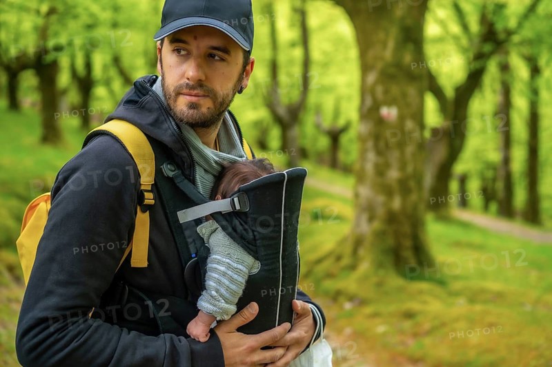 A young father with a yellow backpack and a black cap walking with the newborn child in the backpack on a path in the woods