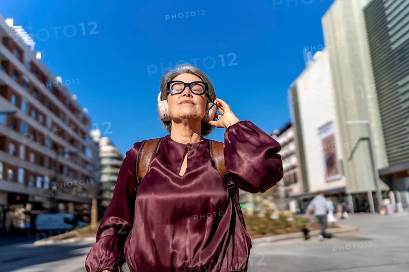 Grey haired woman with eyeglasses and headphones standing outdoors, enjoying music with eyes closed while surrounded by urban buildings and a clear blue sky