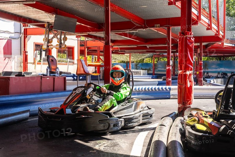 Young driver wearing a bright green suit and an orange helmet, sitting in a go kart on the track, preparing to start a race during a fun day at the outdoor circuit