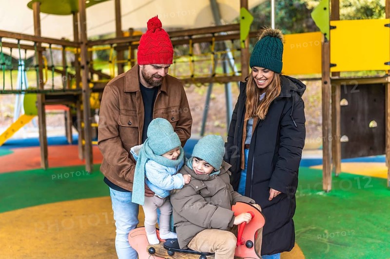 Smiling parents watching their children playing on a rocking horse at the playground during a cold day