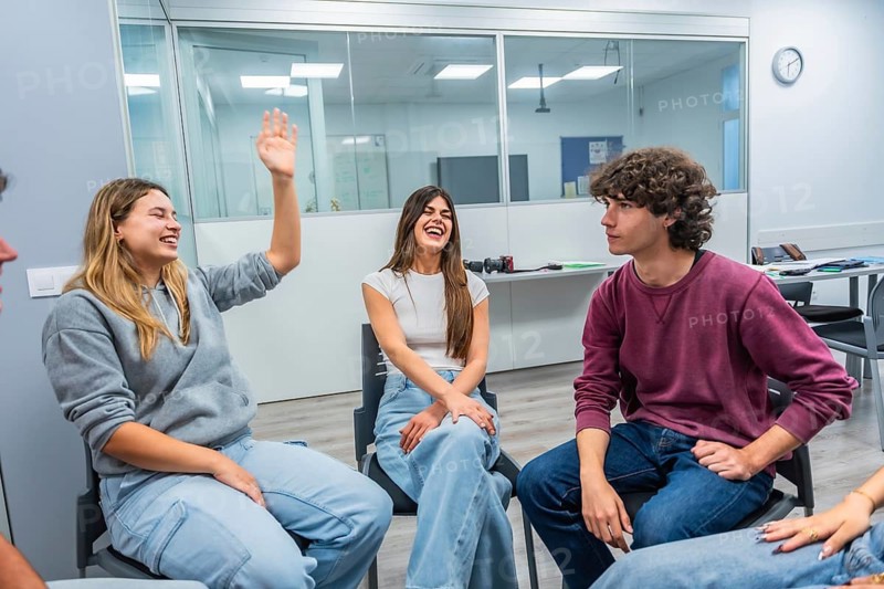 Young university students in a modern classroom laugh, raise hands and share ideas during an interactive group discussion, fostering teamwork, communication and learning