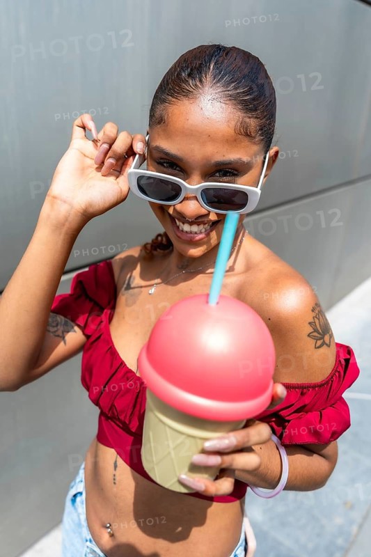 Diverse young woman smiling and holding a novelty ice cream cup with a straw while posing outdoors on a sunny summer day, embodying a cheerful urban lifestyle