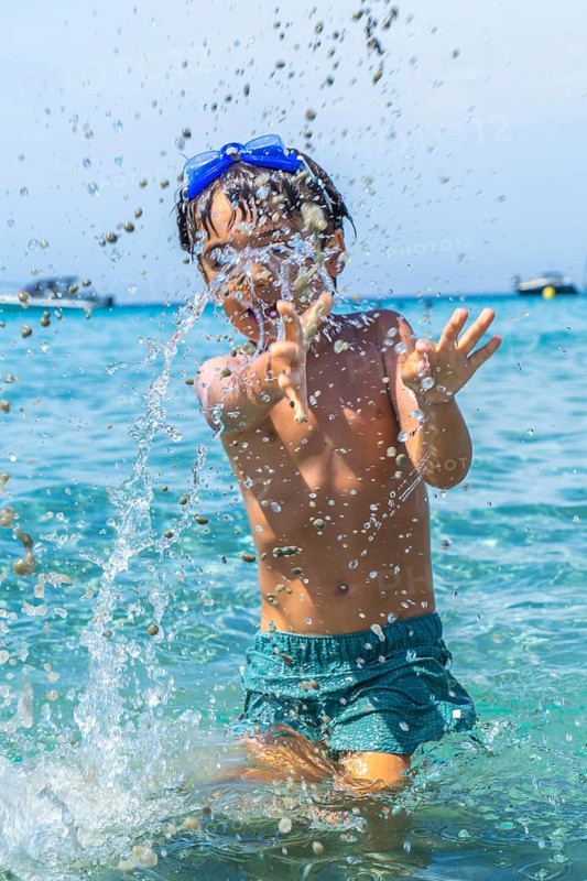Joyful boy wearing blue swimming goggles and green shorts, playing and splashing water in the clear turquoise sea on tabarca island, enjoying summer vacation
