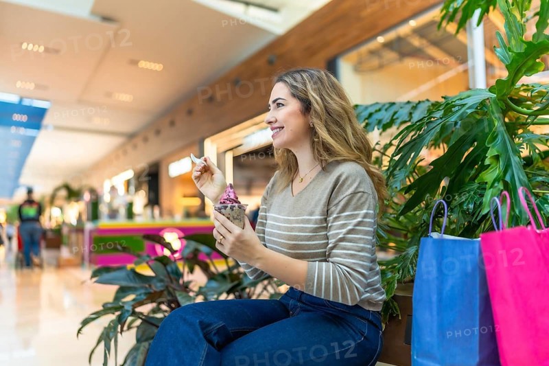 Young woman relaxing amid shopping bags in a bright modern mall, happily enjoying frozen yogurt during a casual retail break, smiling and savoring a sweet treat