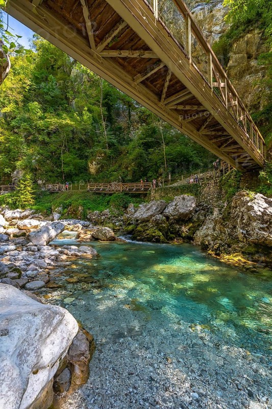 Tourists crossing a wooden footbridge over a clear turquoise river in tolmin gorge, slovenia, surrounded by lush green forest