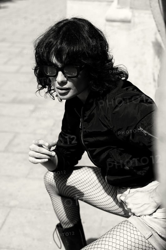 A stylish woman with messy hair and sunglasses exudes confidence in this high-contrast black and white portrait. Dressed in fishnet tights and a bomber jacket, she embodies bold, urban street style