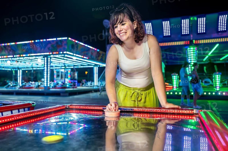 Happy young woman laughing while playing air hockey at a vibrant night amusement park, enjoying fun and entertainment during summer holidays