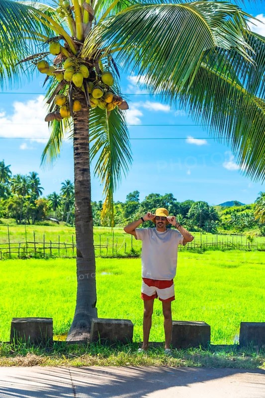 Young man wearing a hat and summer clothes enjoying a sunny day in coron island, philippines, with coconut palm and rice field