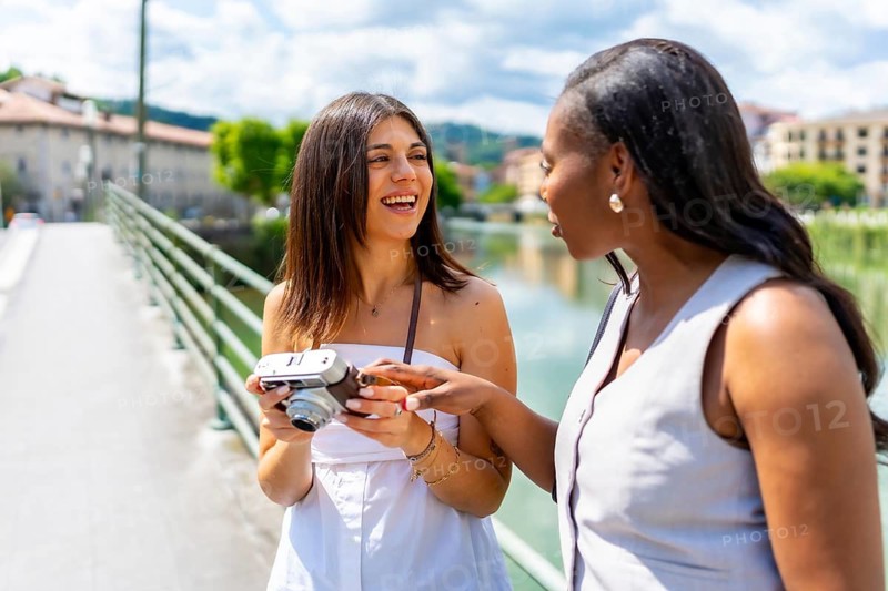 Two happy female tourists using a vintage camera while walking on a bridge in oloron sainte marie, france, during a sunny summer day