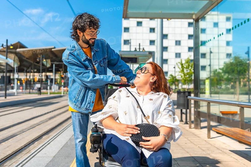Young man joyfully pushing a smiling woman in a wheelchair at a modern tram station, embodying support and inclusion in urban travel