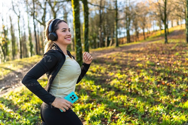 Woman enjoying a morning run through a natural park setting, listening to music with headphones and holding a smartphone, actively pursuing a healthy lifestyle and physical well being outdoors