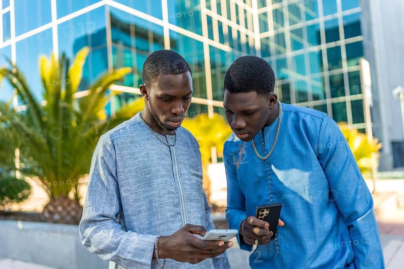 Two senegalese businessmen in traditional dashiki attire using smartphones in front of a modern office building, blending tradition with technology