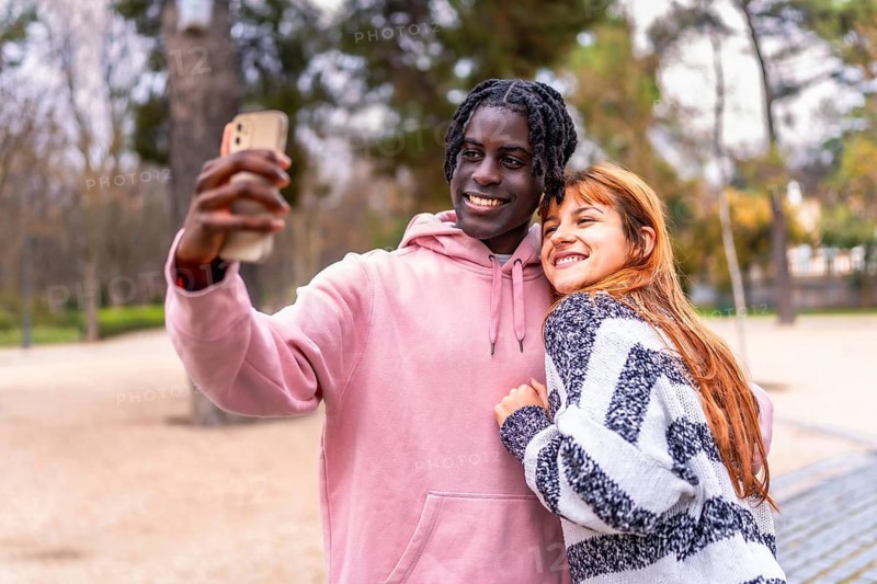 Young multiracial couple taking a selfie with a smartphone in a park, smiling and hugging