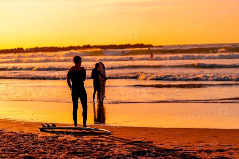 A man stands on the beach with his surfboard, watching the waves. The sky is orange and the water is calm