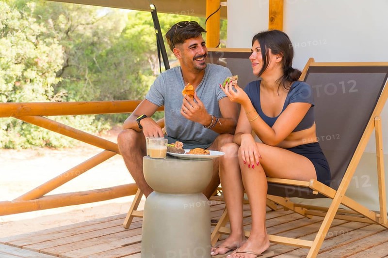 Young couple smiling and having breakfast on the wooden terrace of a glamping tent, surrounded by nature, enjoying a relaxing vacation together