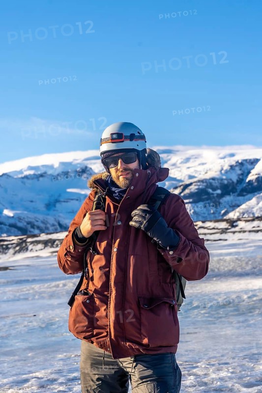A man wearing a red jacket and a helmet stands in the snow. He is holding a camera and has a backpack on his back. The scene is set in a snowy mountain area, with the man posing for a photo