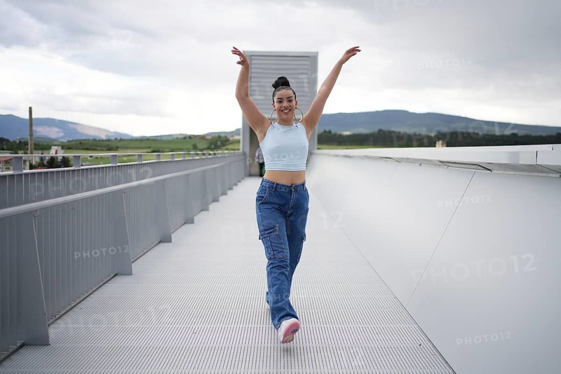 A woman in blue jeans and a blue top is dancing on a bridge. The bridge is made of metal and is located in a mountainous area. The woman is happy and enjoying herself as she dances