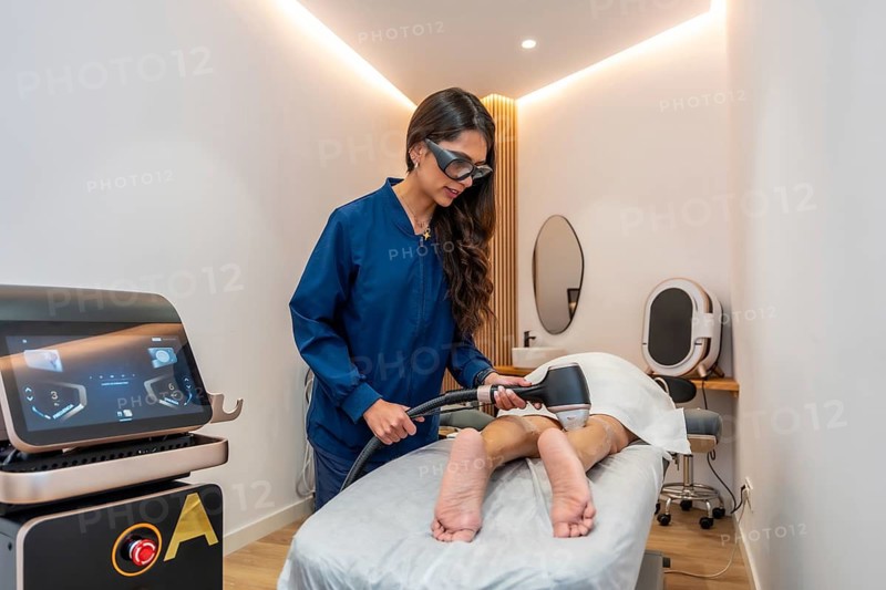 Professional beautician in protective glasses performing laser hair removal on a client's leg, using modern equipment in a clean, modern aesthetic clinic setting