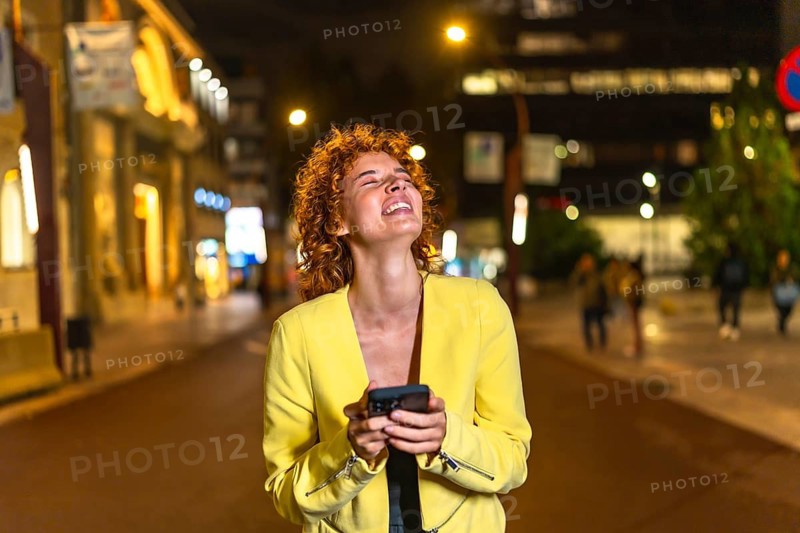Happy woman looking up while strolling along the city streets using mobile phone at night