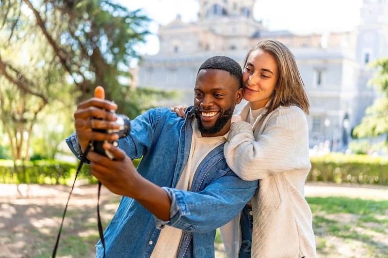 Multiracial couple taking selfie with camera in a park with city views