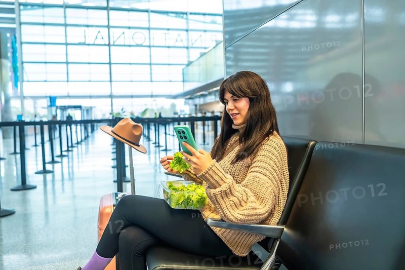 Young woman sitting in a modern airport terminal, eating a fresh green salad from a plastic container and interacting with her mobile phone while waiting for her flight