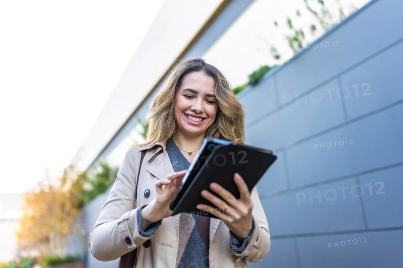 Young businesswoman with a trench coat looking happy while interacting with a digital tablet outdoors, symbolizing connectivity, professional work, and modern technology in an urban environment