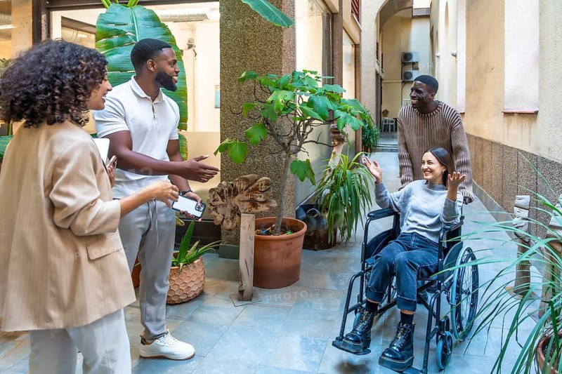 Coworkers eagerly wait for a disabled woman in a wheelchair pushed by an african man
