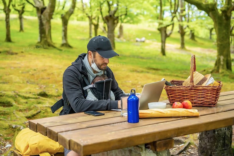 A young Caucasian father teleworking sitting at a picnic table with computer in the woods. Summer lifestyle