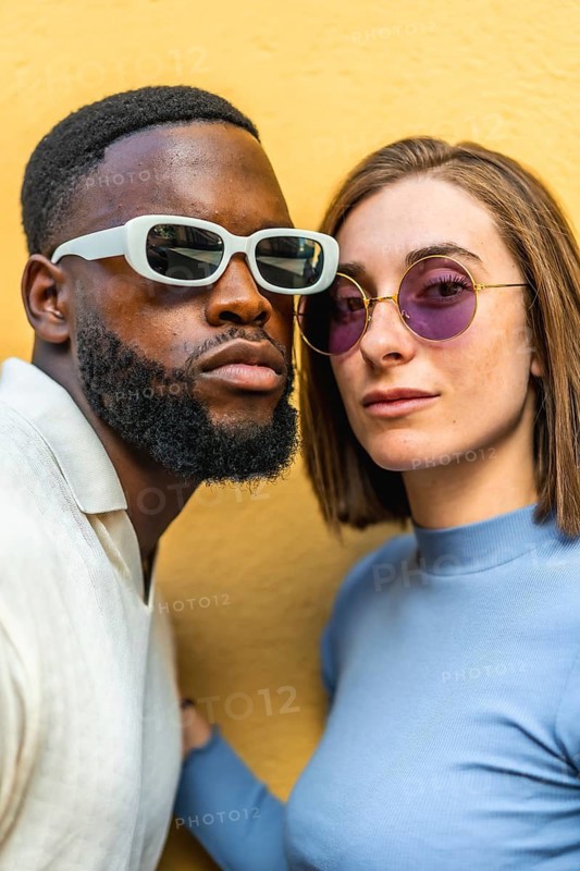Vertical close-up photo of a stylish multiracial couple wearing sunglasses looking at camera together against yellow background