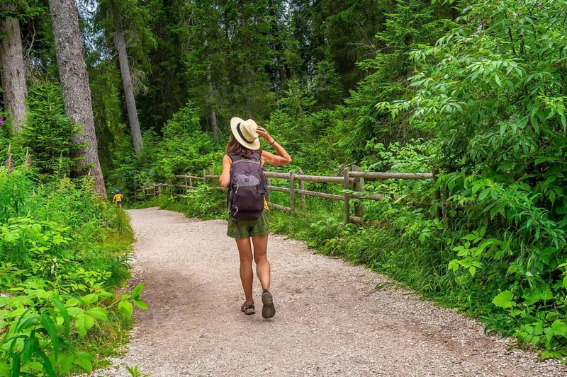 Female tourist with straw hat and backpack walking on gravel path in forest near lake carezza, dolomites, italy, enjoying summer vacation