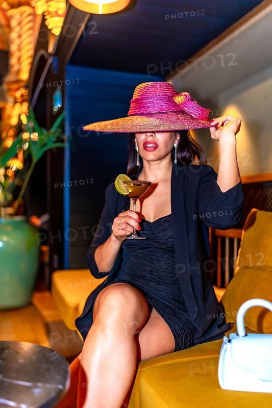 Vertical photo of a woman wearing elegant clothes and hat sitting drinking luxury cocktail in a night pub