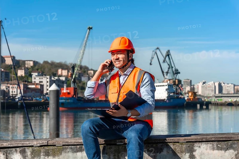 Young male port worker wearing safety helmet and vest, holding clipboard and talking on a mobile phone while sitting by the harbor with cargo ships and cranes in the background