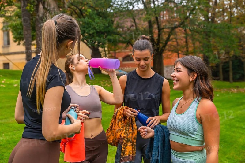 Four young women taking a break and hydrating with reusable water bottles, conversing and smiling in a green park after exercising, fostering friendship and healthy activity