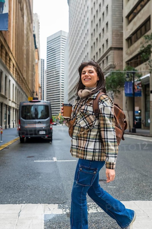 Young woman smiling while walking across a san francisco city street, holding a disposable coffee cup and carrying a backpack, with tall buildings and a van in the background