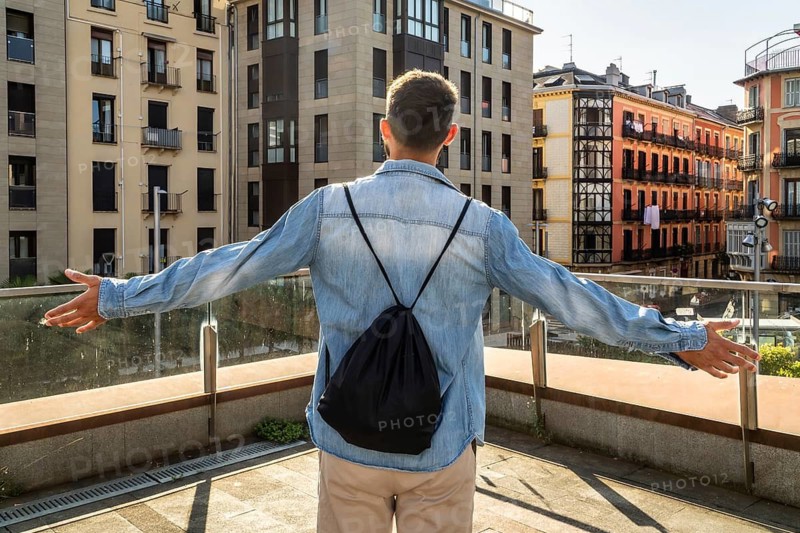 Man with backpack standing on a terrace with arms outstretched, experiencing freedom and success while overlooking a vibrant urban cityscape under a bright sky