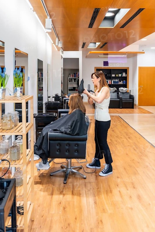 Stylist blow drying a female client's hair, providing a professional beauty treatment in a brightly lit, modern hairdressing salon with wooden floors and contemporary design elements