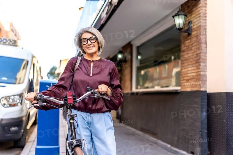 Senior woman with gray hair and glasses smiling at the camera while walking a bicycle on a urban street, embracing an active and healthy lifestyle outdoors