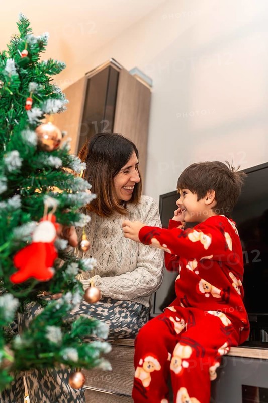 Mother and son in pajamas laughing as they decorate a small christmas tree at home, sharing cozy, festive moments and holiday traditions together in a warm, joyful scene