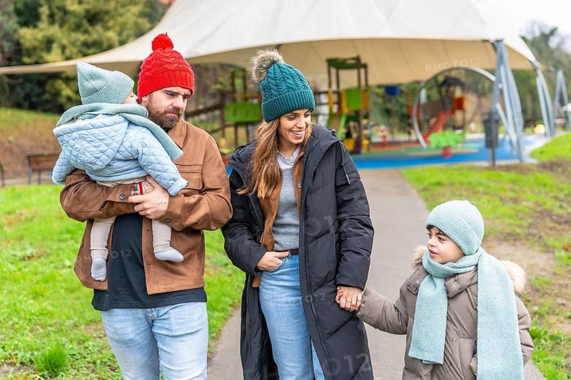 Smiling parents holding their children's hands are walking near a playground on a cold winter day